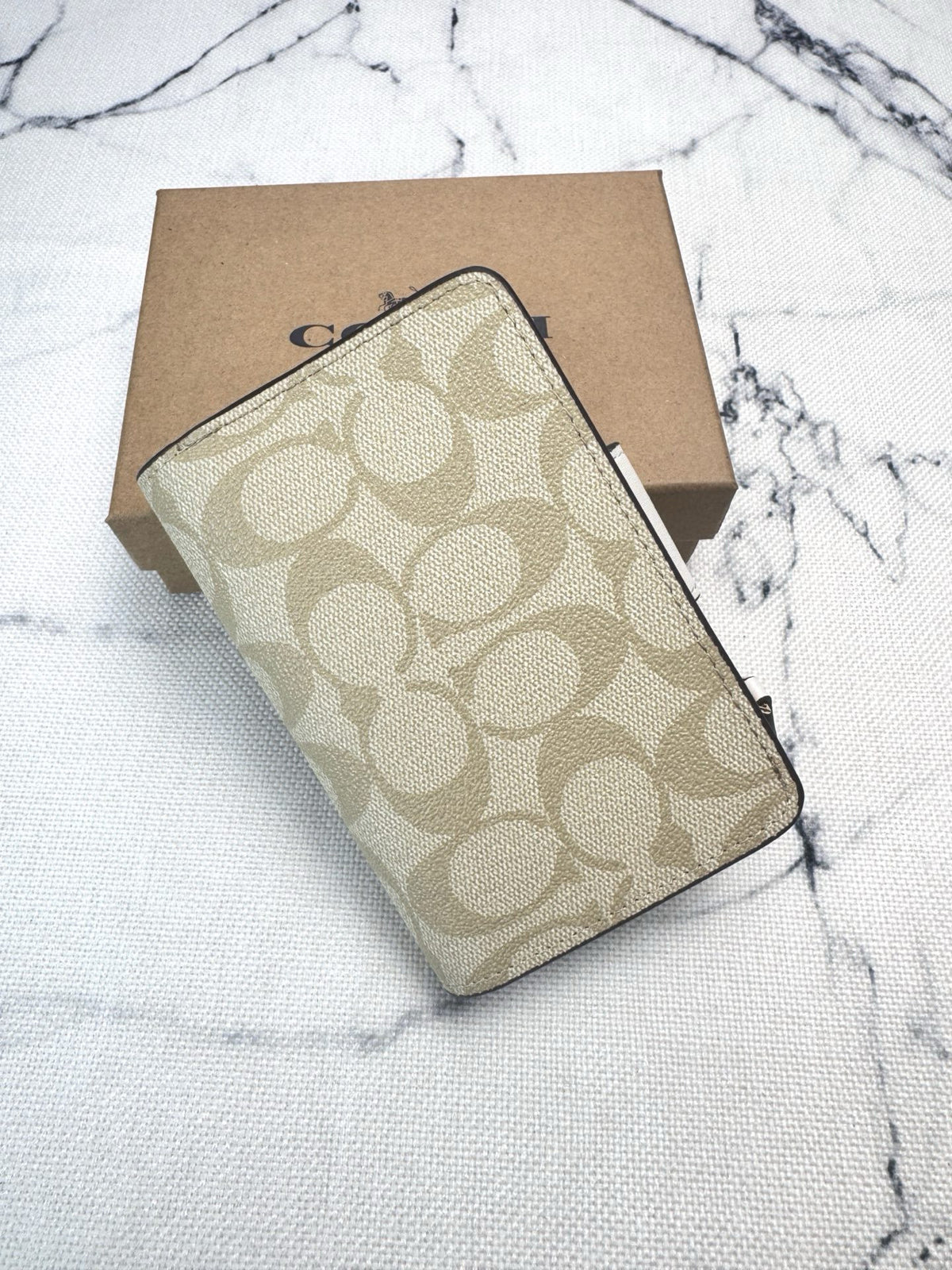 A Coach signature canvas zip-around wallet in beige and brown, shown with its brown cardboard packaging box on a white marble-patterned surface.