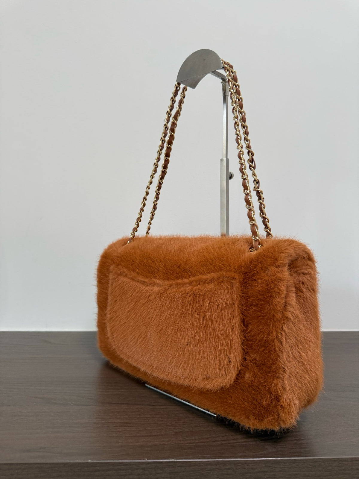 A brown faux fur shoulder bag with a chain strap, displayed on a stand on a dark wooden surface against a light gray background.