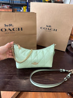 A Coach shoulder bag in pale mint green quilted leather with gold hardware, held by a hand in front of two brown Coach New York paper shopping bags on a dark wooden table.