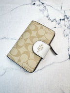A Coach zip-around wallet in beige signature monogram canvas with a white leather tab and gold logo, placed on a white marble-patterned surface.