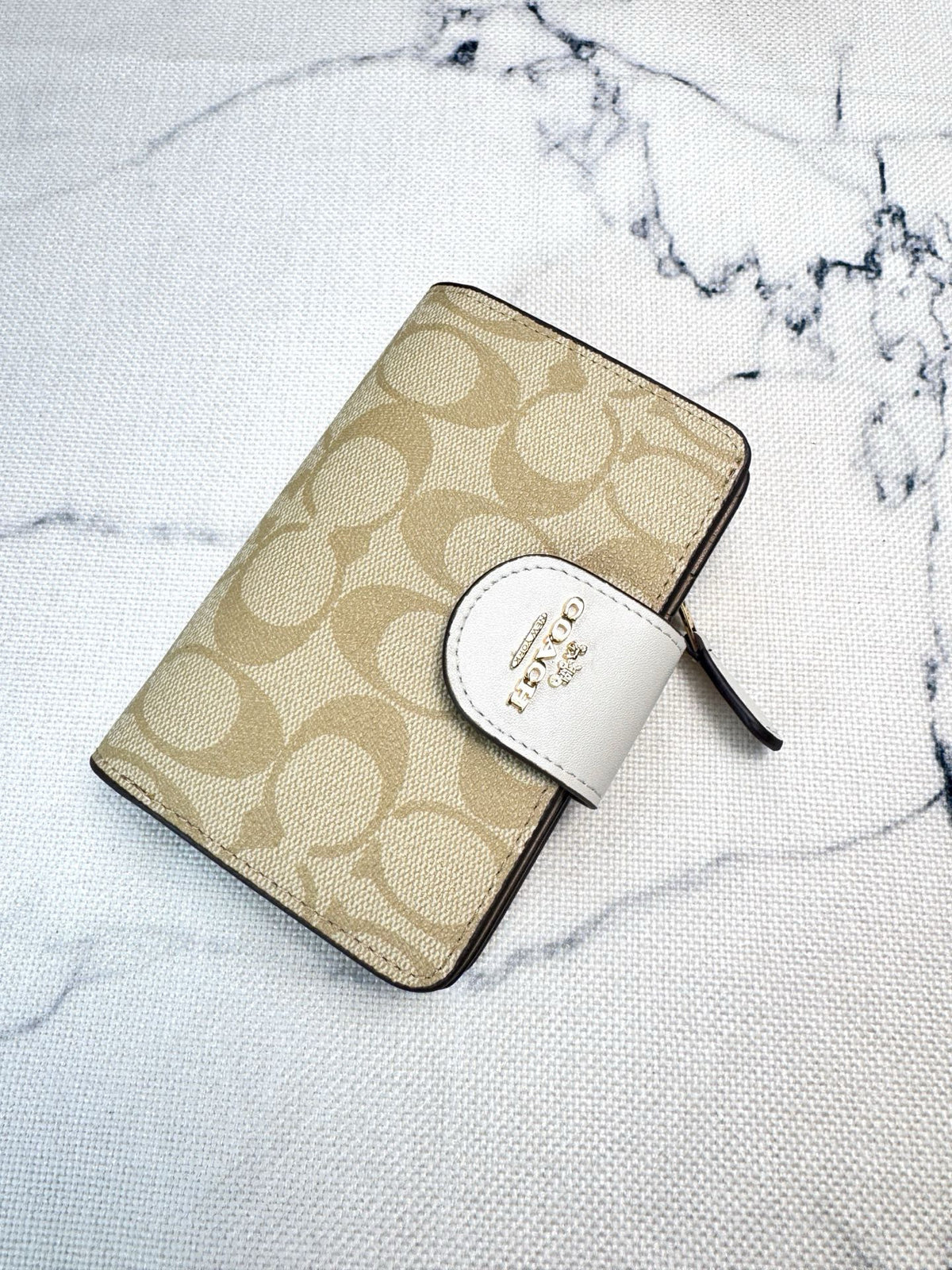 A Coach zip-around wallet in beige signature monogram canvas with a white leather tab and gold logo, placed on a white marble-patterned surface.