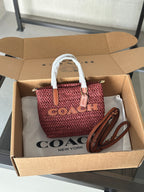A Coach woven straw handbag in dusty rose pink with tan leather trim and white handles, displayed inside an open brown cardboard box with Coach New York branding on the tissue paper.