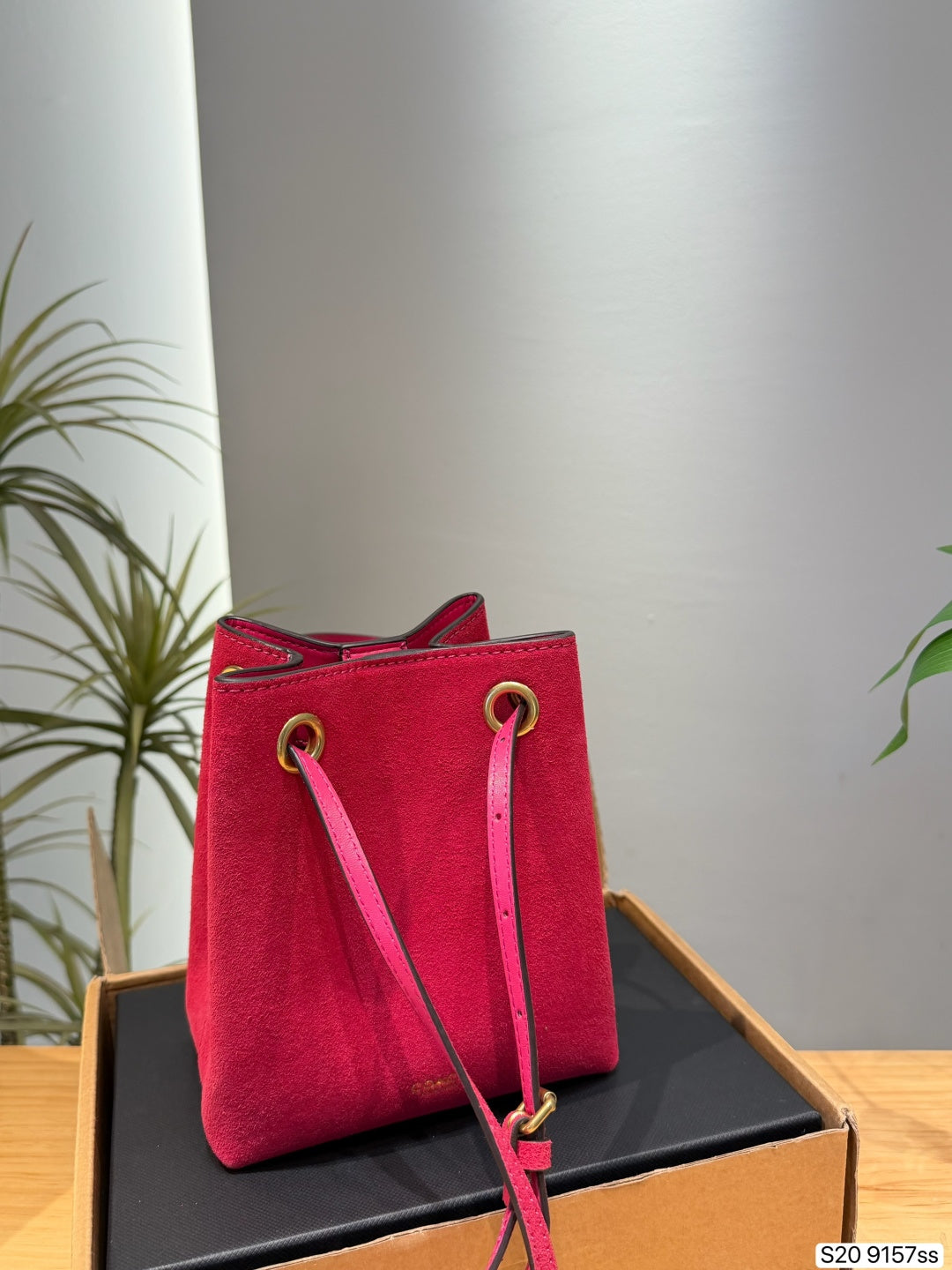 A bright pink-red suede bucket bag with gold-tone hardware, displayed on a black surface inside a cardboard box. The background features a neutral gray wall and a green plant on the left.