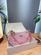 A pink Coach handbag with a signature monogram pattern, gold-tone chain strap, and pink flower charms, displayed inside an open white Coach box on a wooden table.