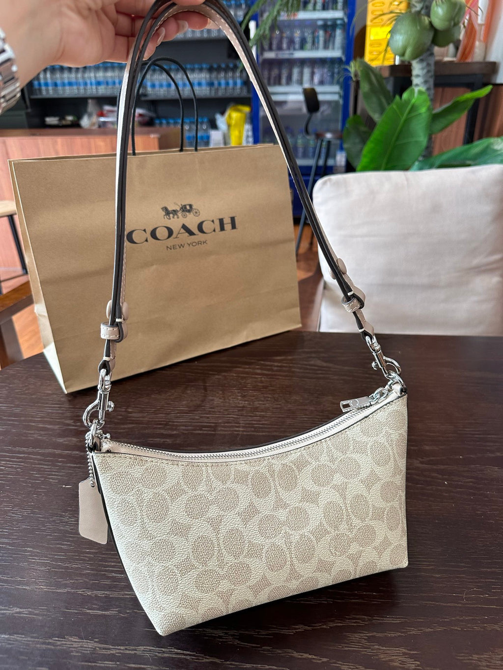 A Coach shoulder bag in beige signature monogram canvas with silver-tone hardware, held by a hand in front of a Coach paper shopping bag on a dark wooden table.