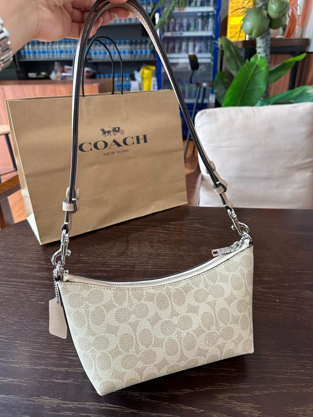 A Coach shoulder bag in beige signature monogram canvas with silver-tone hardware, held by a hand in front of a Coach paper shopping bag on a dark wooden table.
