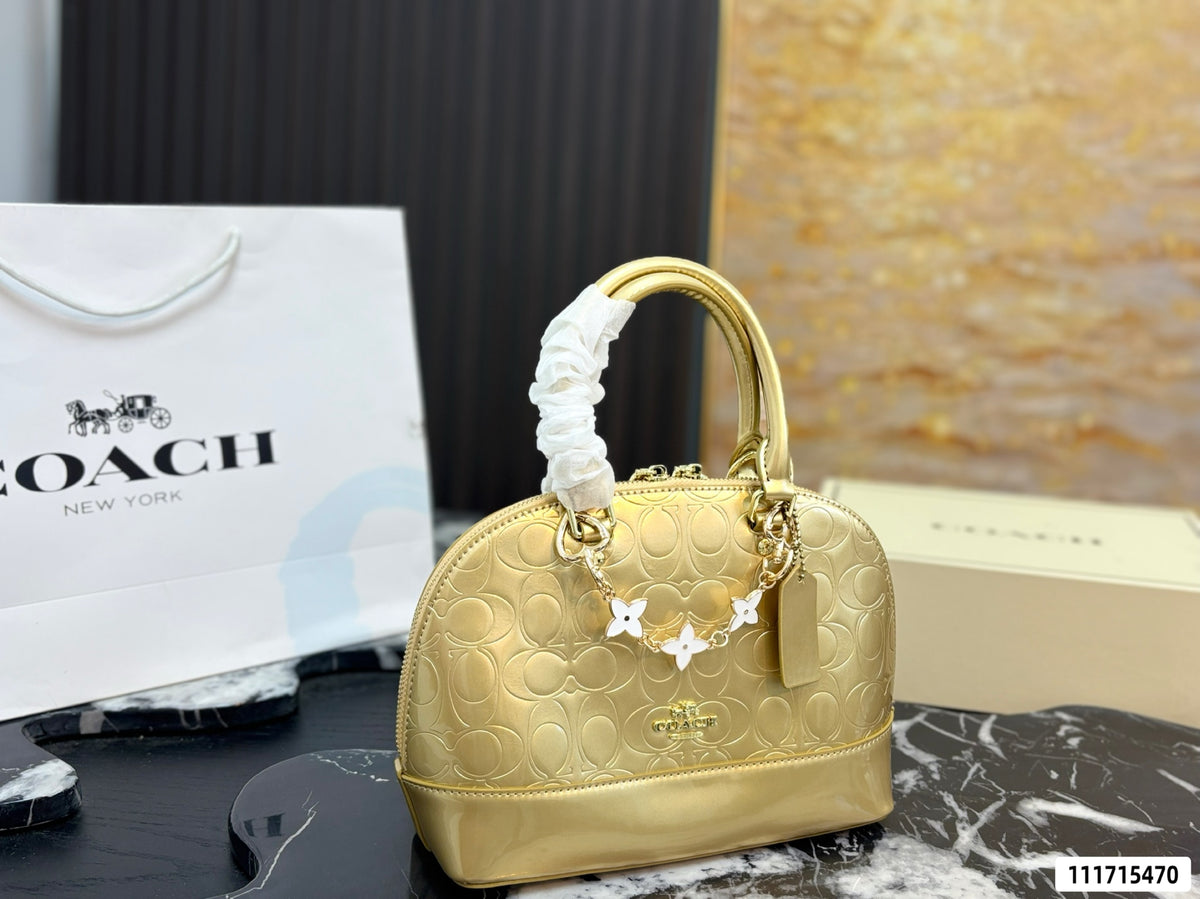 A gold Coach handbag with a white ruched handle and white floral charms, displayed next to a Coach shopping bag and a Coach box on a marble surface.