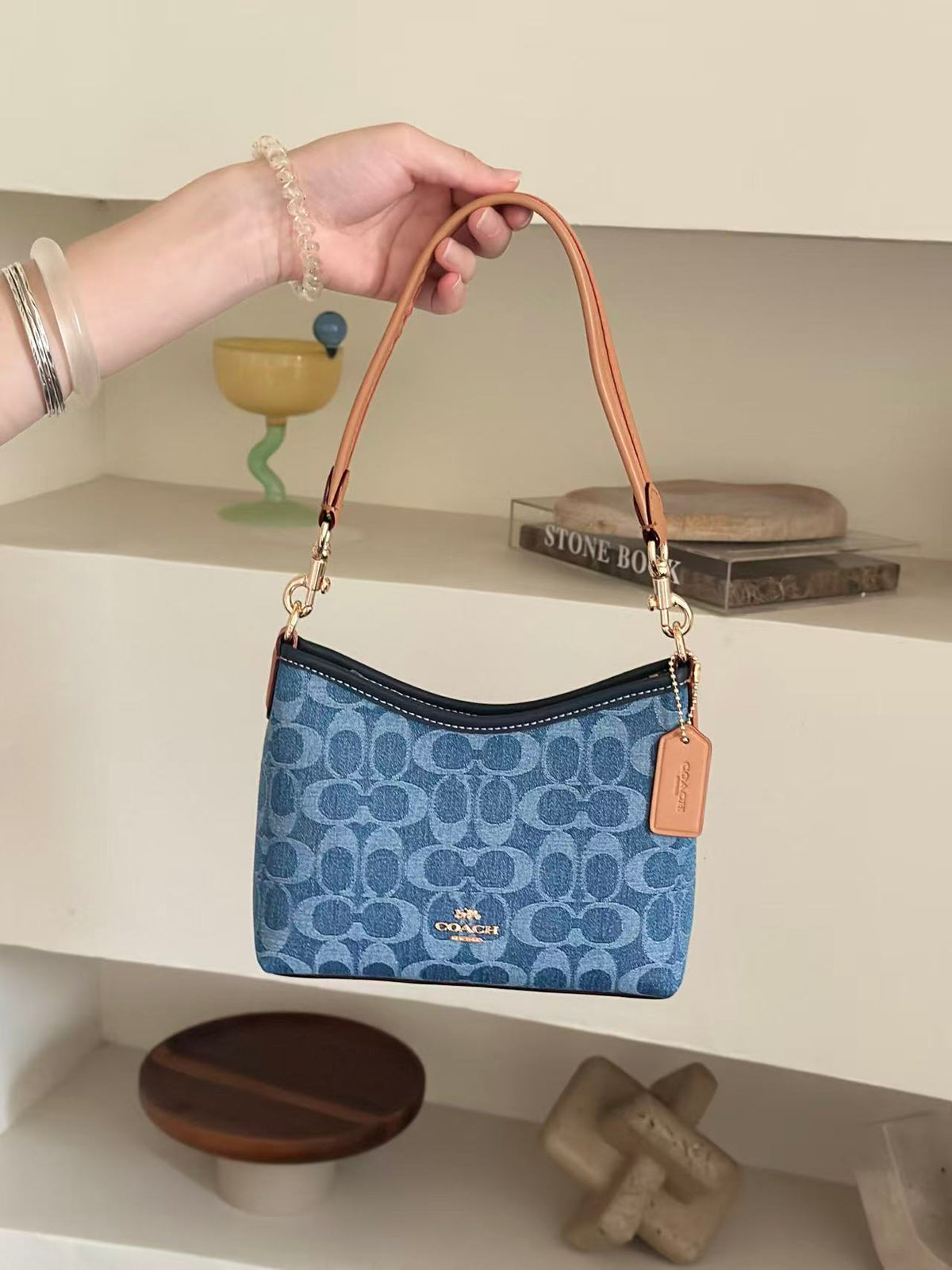 A blue patterned Coach shoulder bag with tan leather trim and strap is held by a person's hand against a beige shelving background.