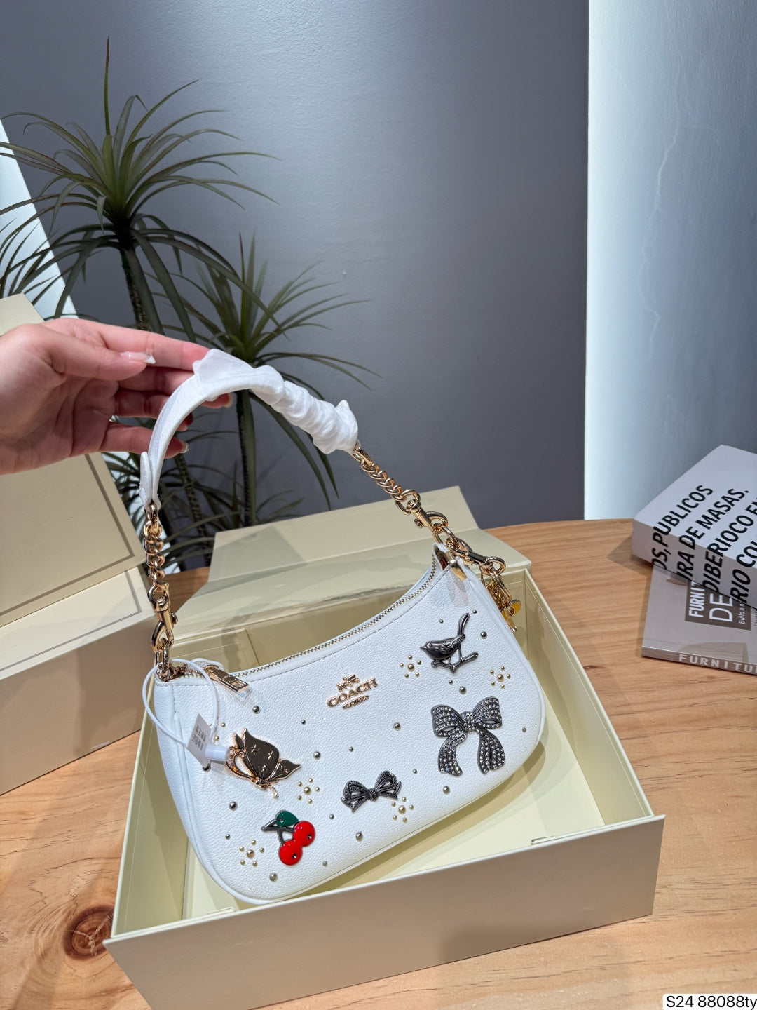 A white Coach handbag with black bow and cherry embellishments, held by a hand over an open cream-colored Coach box on a wooden table.