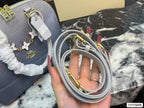 A hand holding a light gray leather shoulder strap with gold-tone hardware, next to a matching gray quilted handbag with a white ruffled handle and butterfly charm on a black-and-white marble surface.