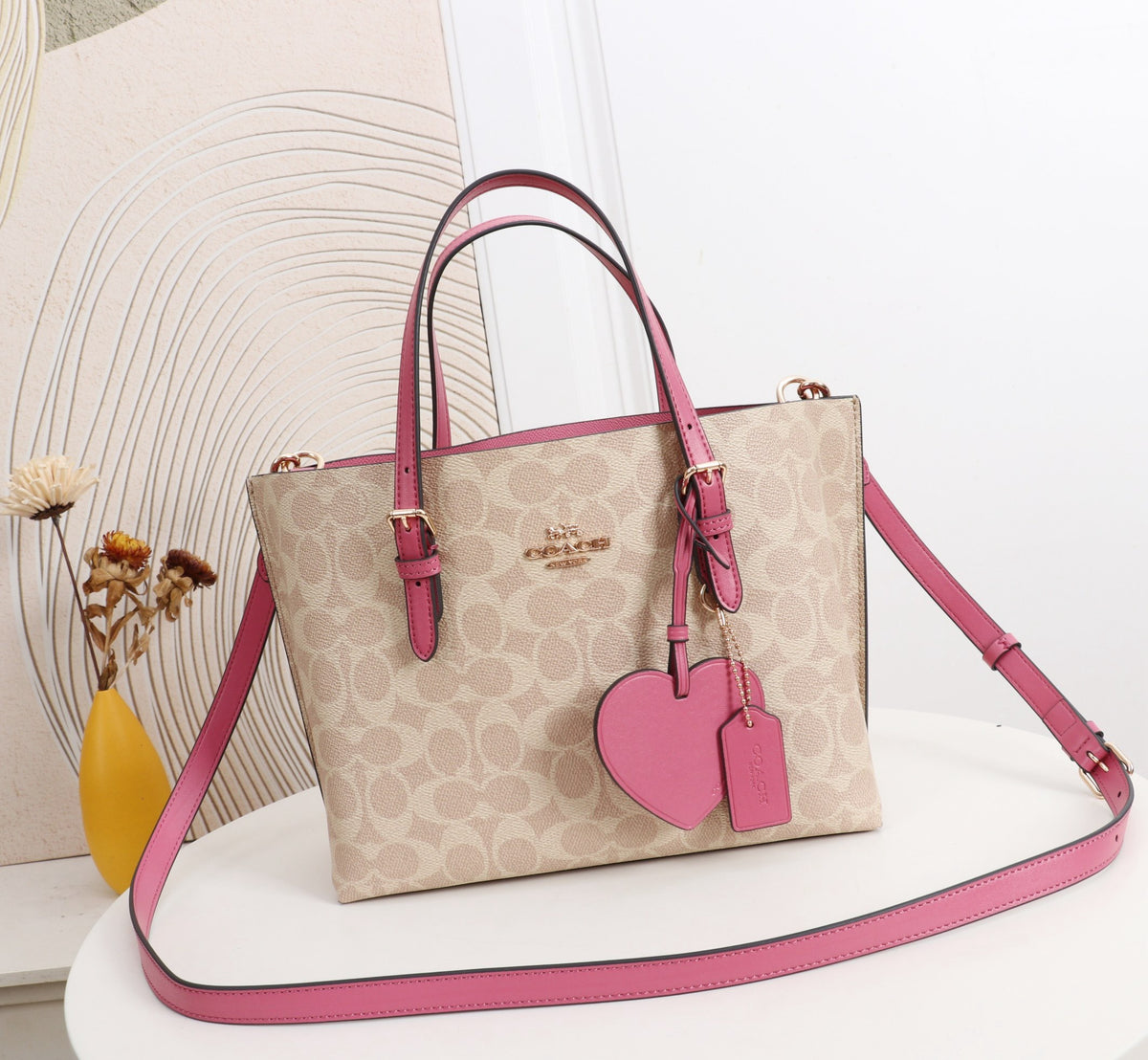 A beige and pink Coach signature monogram tote bag with pink leather handles, a matching pink heart-shaped charm, and a long pink shoulder strap, displayed on a white table with a decorative background.