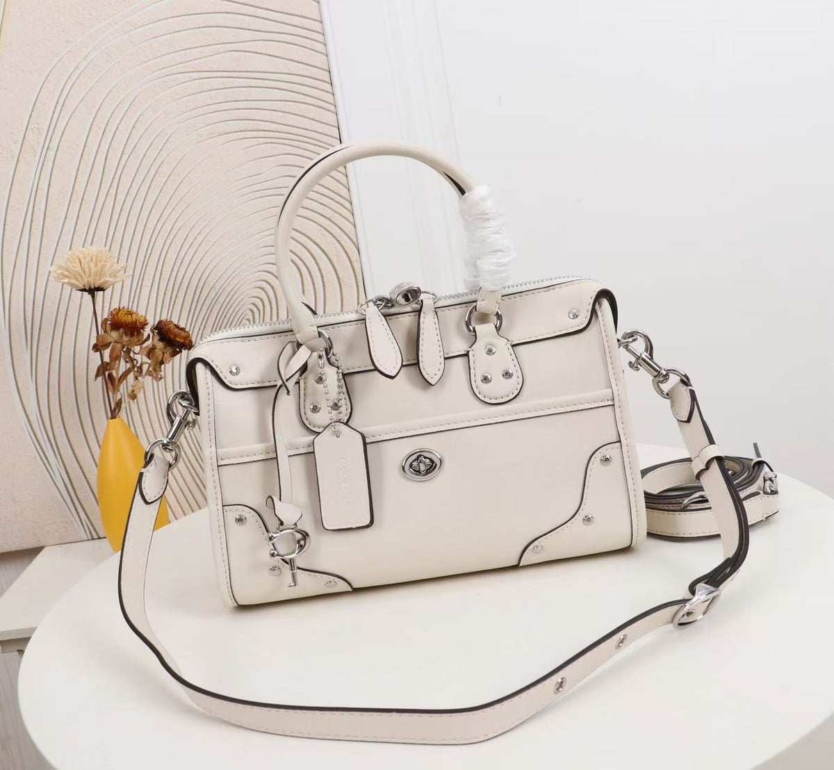 An off-white leather handbag with silver-tone hardware, a top handle, and a detachable shoulder strap, displayed on a white table with a decorative background.