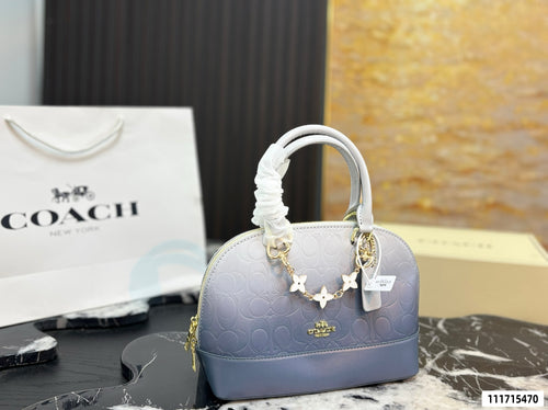 A Coach handbag in a dusty lavender-blue color, featuring the brand's signature monogram pattern, a white fabric-wrapped top handle, and a gold-tone star charm. A white Coach shopping bag is visible in the background on a black marble surface.