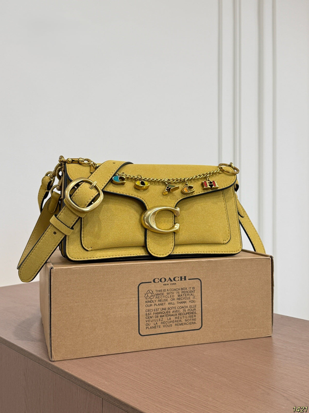 A Coach shoulder bag in mustard yellow leather with gold hardware and decorative charms, displayed on top of a brown Coach box against a white paneled wall.
