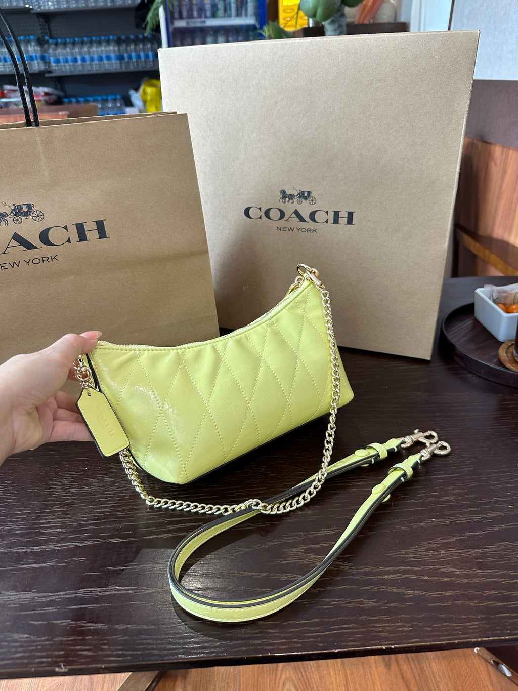 A Coach handbag in light yellow quilted leather with a gold chain strap and matching wristlet strap, displayed on a dark wooden table with Coach shopping bags in the background.