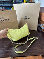 A Coach handbag in light yellow quilted leather with a gold chain strap and matching wristlet strap, displayed on a dark wooden table with Coach shopping bags in the background.