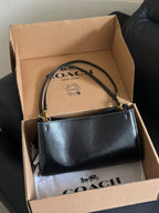 A black Coach handbag with gold-tone hardware resting inside an open brown cardboard Coach box, with the Coach logo visible on the box interior and dust bag.