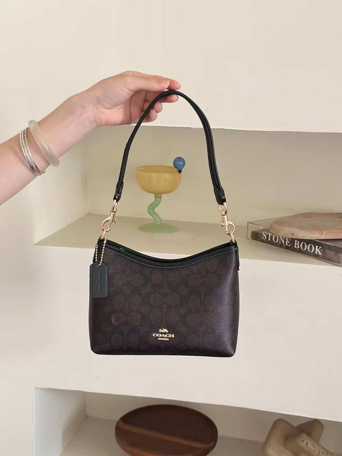 A dark brown Coach hobo bag with a signature monogram pattern, held by a hand against a beige shelf background.