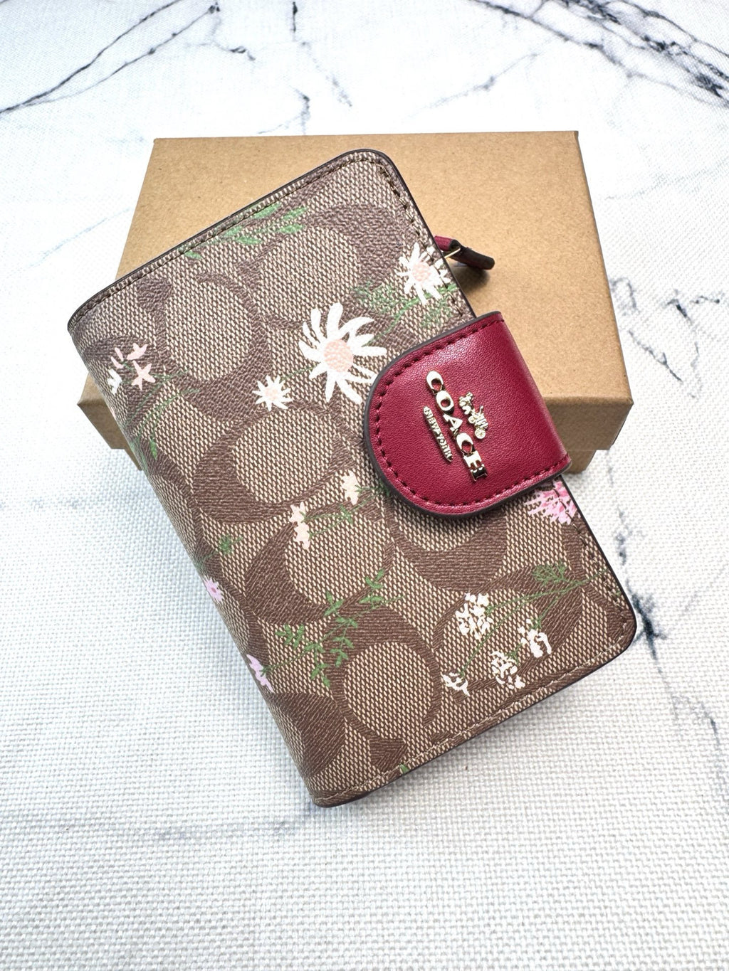 A Coach wallet in the signature brown monogram canvas with white and pink floral motifs, featuring a red leather snap tab with the Coach logo, resting on a brown cardboard box over a white marble surface.