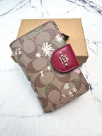 A Coach wallet in the signature brown monogram canvas with white and pink floral motifs, featuring a red leather snap tab with the Coach logo, resting on a brown cardboard box over a white marble surface.