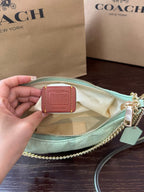 A Coach handbag in pale mint green is shown open with a hand holding a small leather Coach tag inside. The bag has a gold chain strap and zipper, with Coach New York shopping bags in the background on a dark wooden surface.