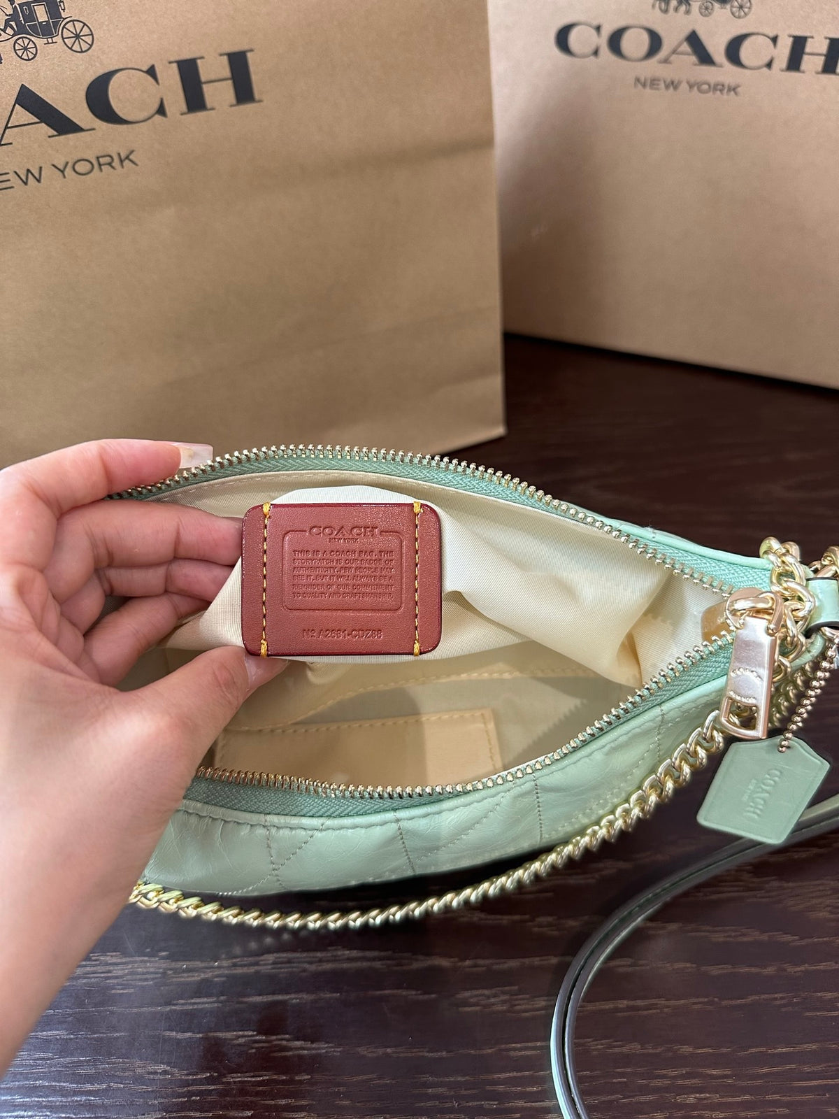 A Coach handbag in pale mint green is shown open with a hand holding a small leather Coach tag inside. The bag has a gold chain strap and zipper, with Coach New York shopping bags in the background on a dark wooden surface.