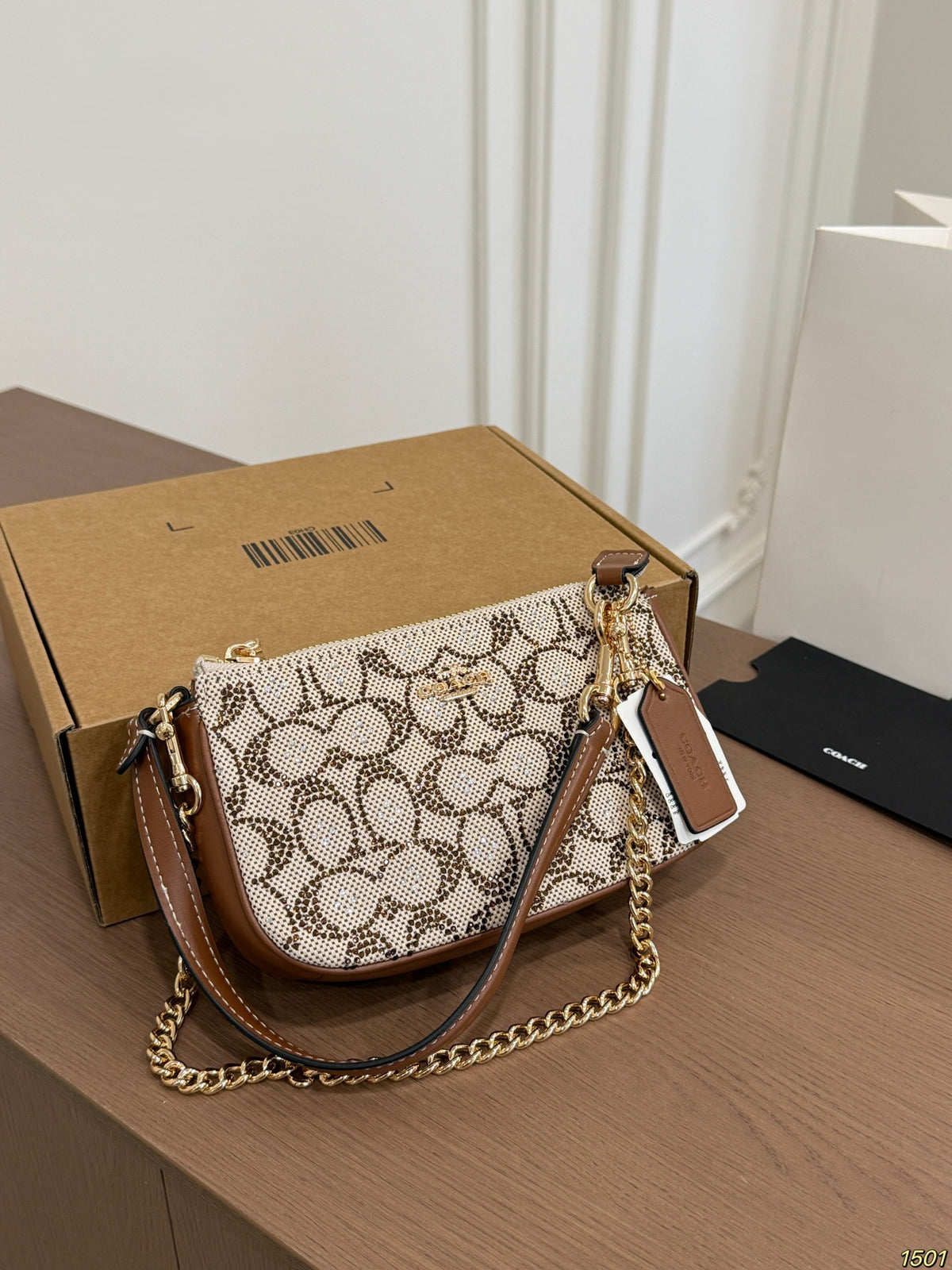 A Coach monogram shoulder bag with brown leather trim and a gold chain strap, resting on a wooden table next to its brown cardboard box.