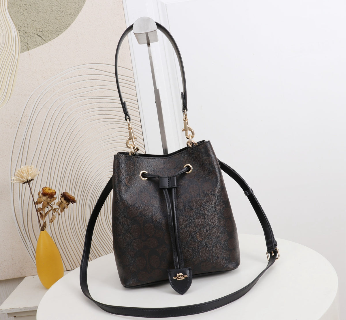 A dark brown monogram bucket bag with a black leather top handle and shoulder strap, displayed on a white round table with a decorative background.