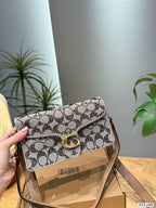 A Coach monogram crossbody bag with a gold-tone C clasp, held by a hand over a cardboard box in an indoor setting with plants in the background.