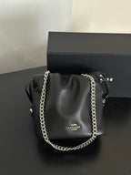 A black Coach shoulder bag with a silver chain strap, displayed in front of its black branded box on a dark surface.