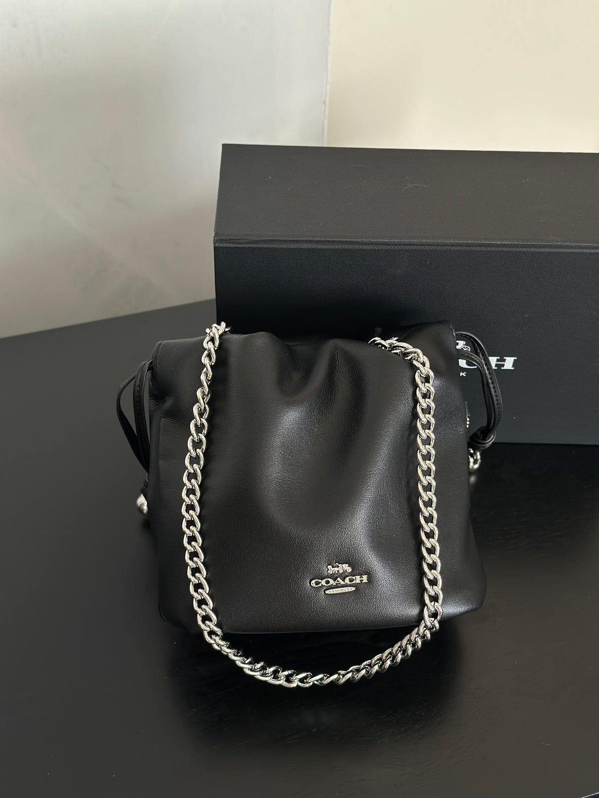 A black Coach shoulder bag with a silver chain strap, displayed in front of its black branded box on a dark surface.