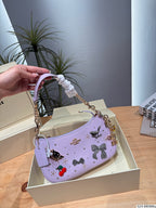 A lavender purple crescent-shaped handbag with decorative bows, rhinestones, and a gold chain handle, held by a hand over an open cream-colored box.