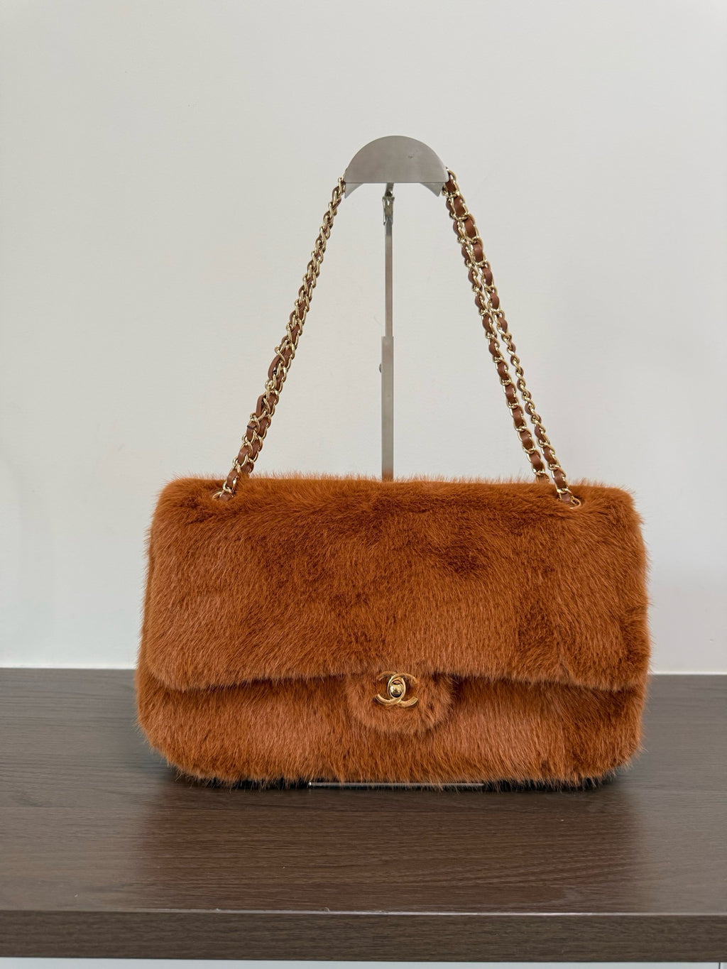 A brown faux fur shoulder bag with a gold-tone interlocking CC clasp and chain strap, displayed on a stand on a dark wooden surface against a white background.