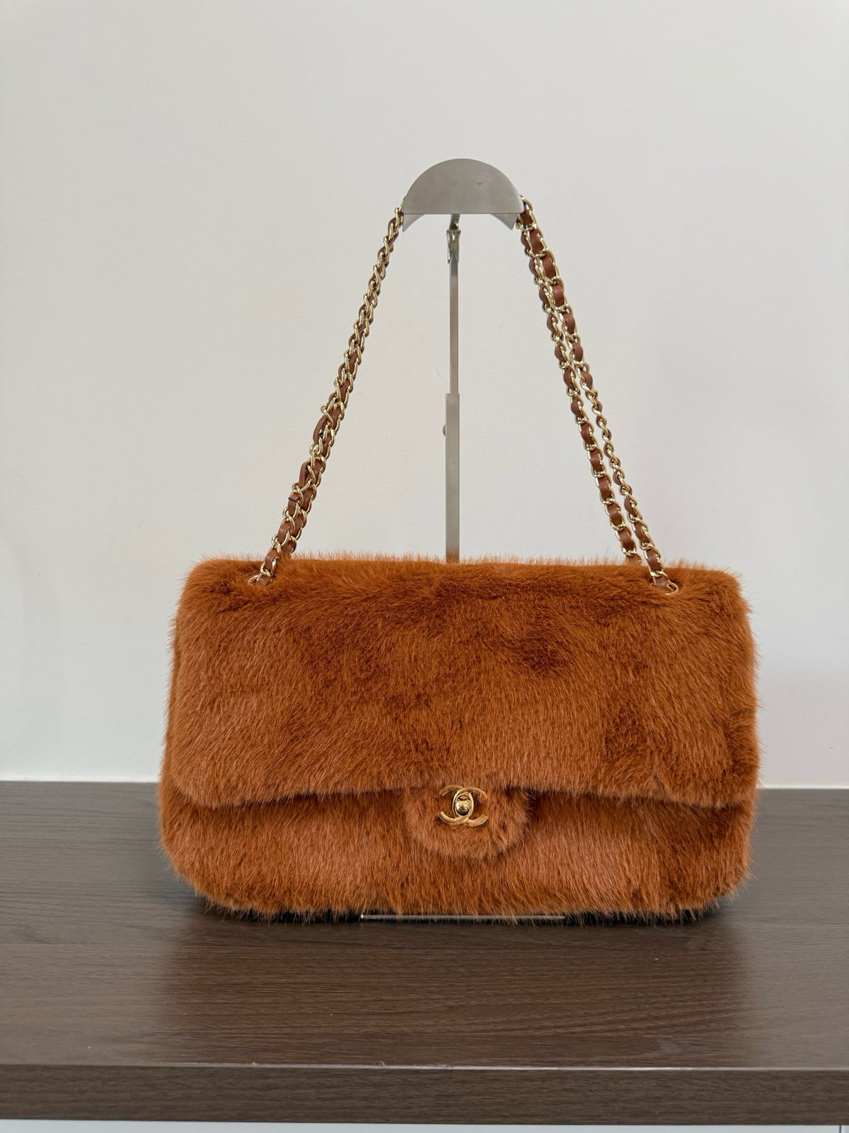 A brown faux fur shoulder bag with a gold-tone interlocking CC clasp and chain strap, displayed on a stand on a dark wooden surface against a white background.