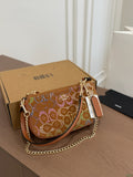A brown leather shoulder bag with a gold chain strap and colorful rhinestone monogram pattern, resting on a wooden table next to its brown cardboard box.
