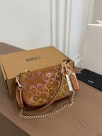 A brown leather shoulder bag with a gold chain strap and colorful rhinestone monogram pattern, resting on a wooden table next to its brown cardboard box.