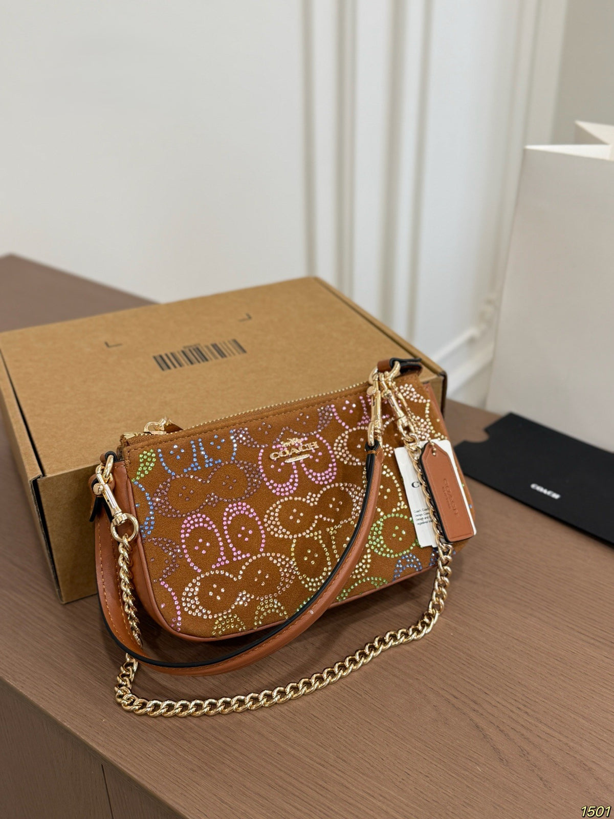 A brown leather shoulder bag with a gold chain strap and colorful rhinestone monogram pattern, resting on a wooden table next to its brown cardboard box.