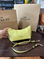 A Coach shoulder bag in light yellow-green patent leather with a quilted diamond pattern, gold-tone chain strap, and a matching detachable wristlet strap, shown on a dark wooden table with Coach shopping bags in the background.