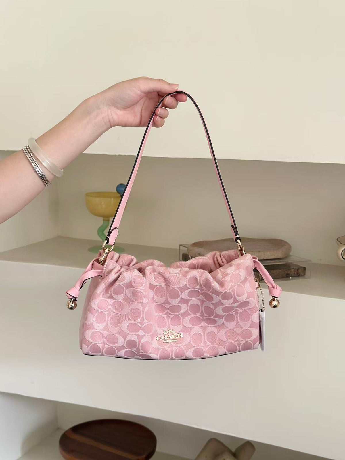 A pink Coach handbag with a signature monogram pattern, held by a person's hand in front of a neutral indoor background.