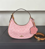 A pink Coach hobo shoulder bag with a signature monogram pattern, tan leather trim, a matching pink flower-shaped charm, and a tan leather strap on a dark gray surface.