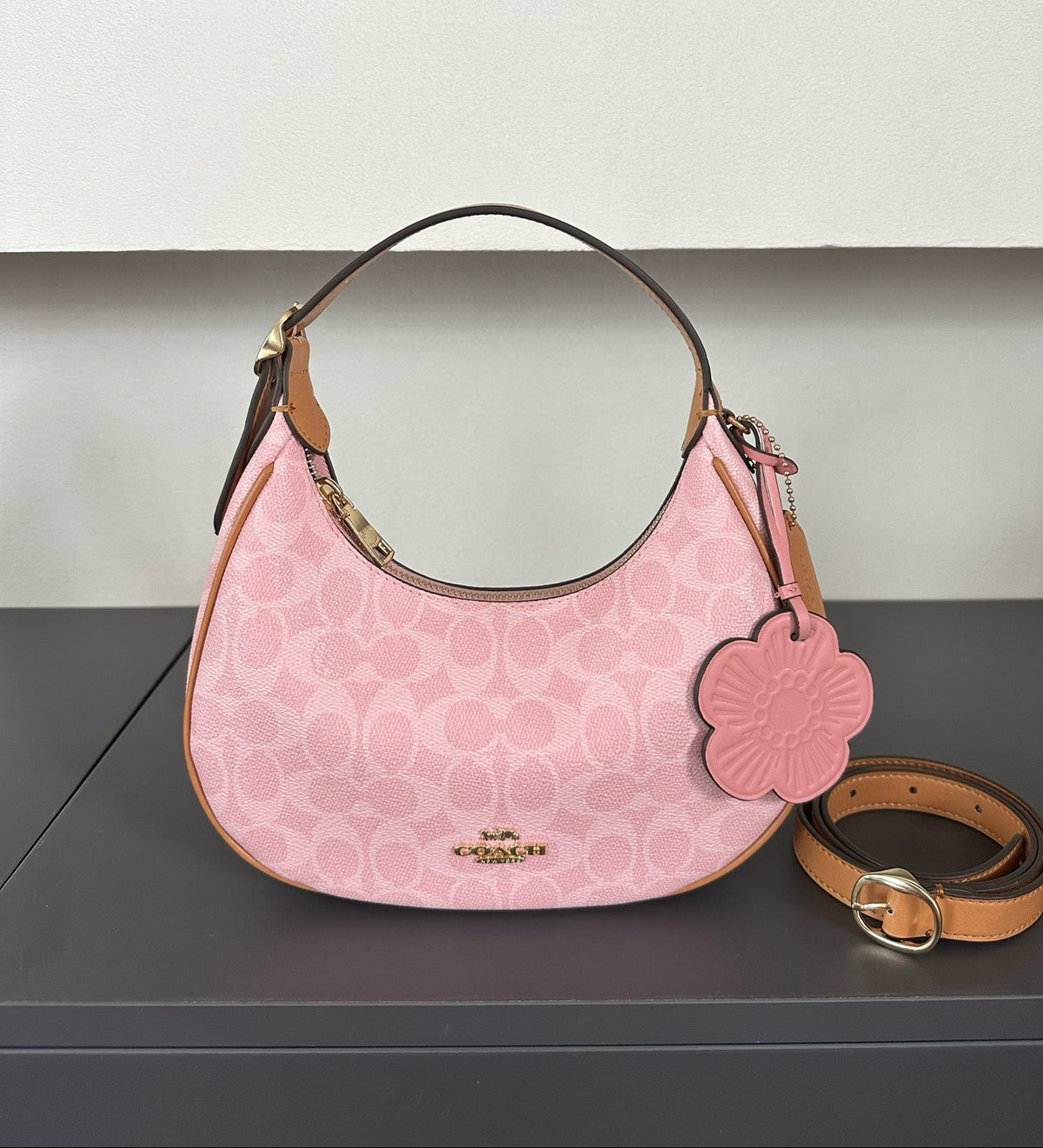 A pink Coach hobo shoulder bag with a signature monogram pattern, tan leather trim, a matching pink flower-shaped charm, and a tan leather strap on a dark gray surface.