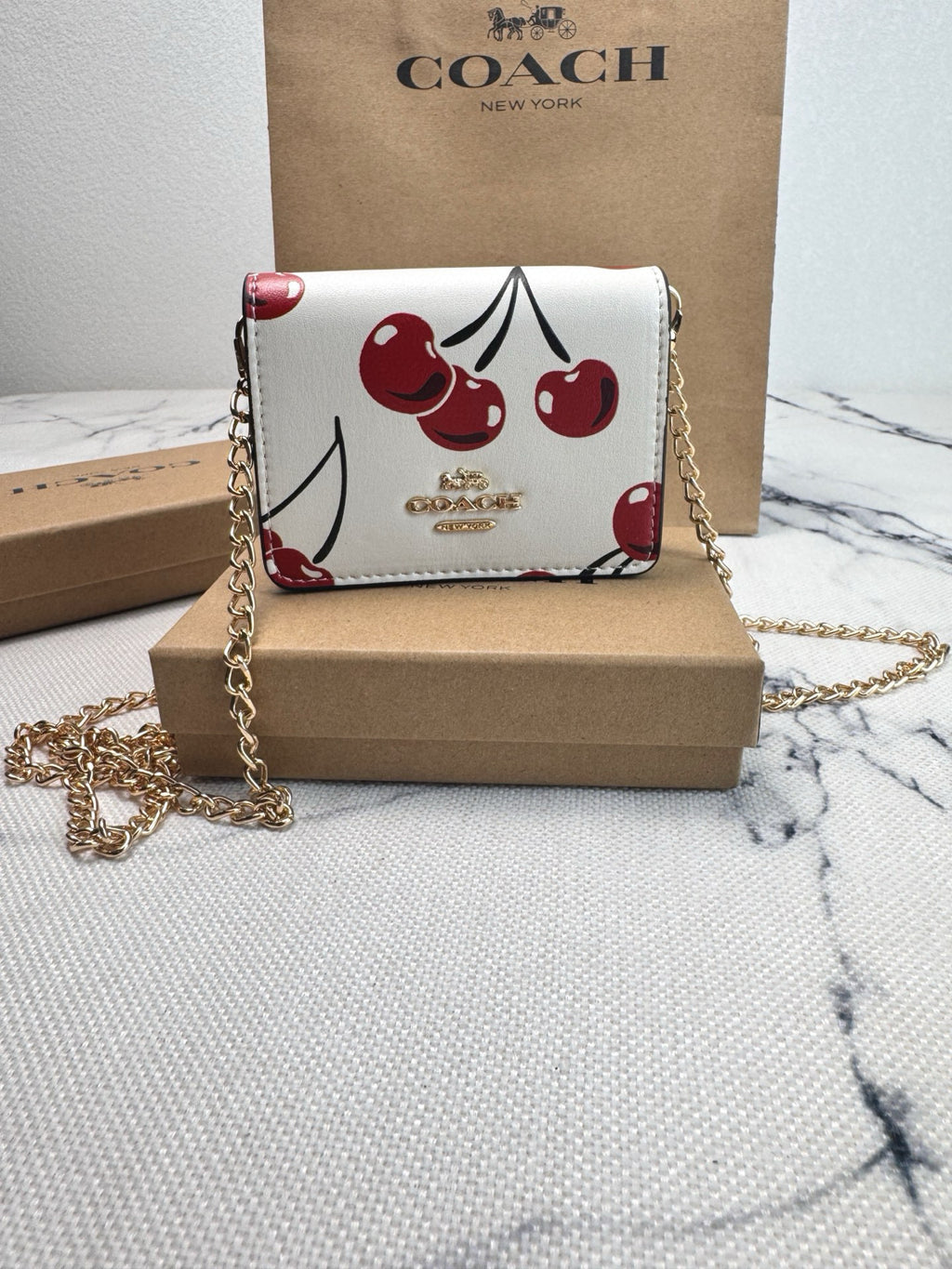 A Coach crossbody bag with a white base and a red cherry print, displayed on a brown cardboard box with its signature Coach box and paper bag in the background.