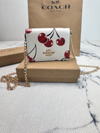 A Coach crossbody bag with a white base and a red cherry print, displayed on a brown cardboard box with its signature Coach box and paper bag in the background.