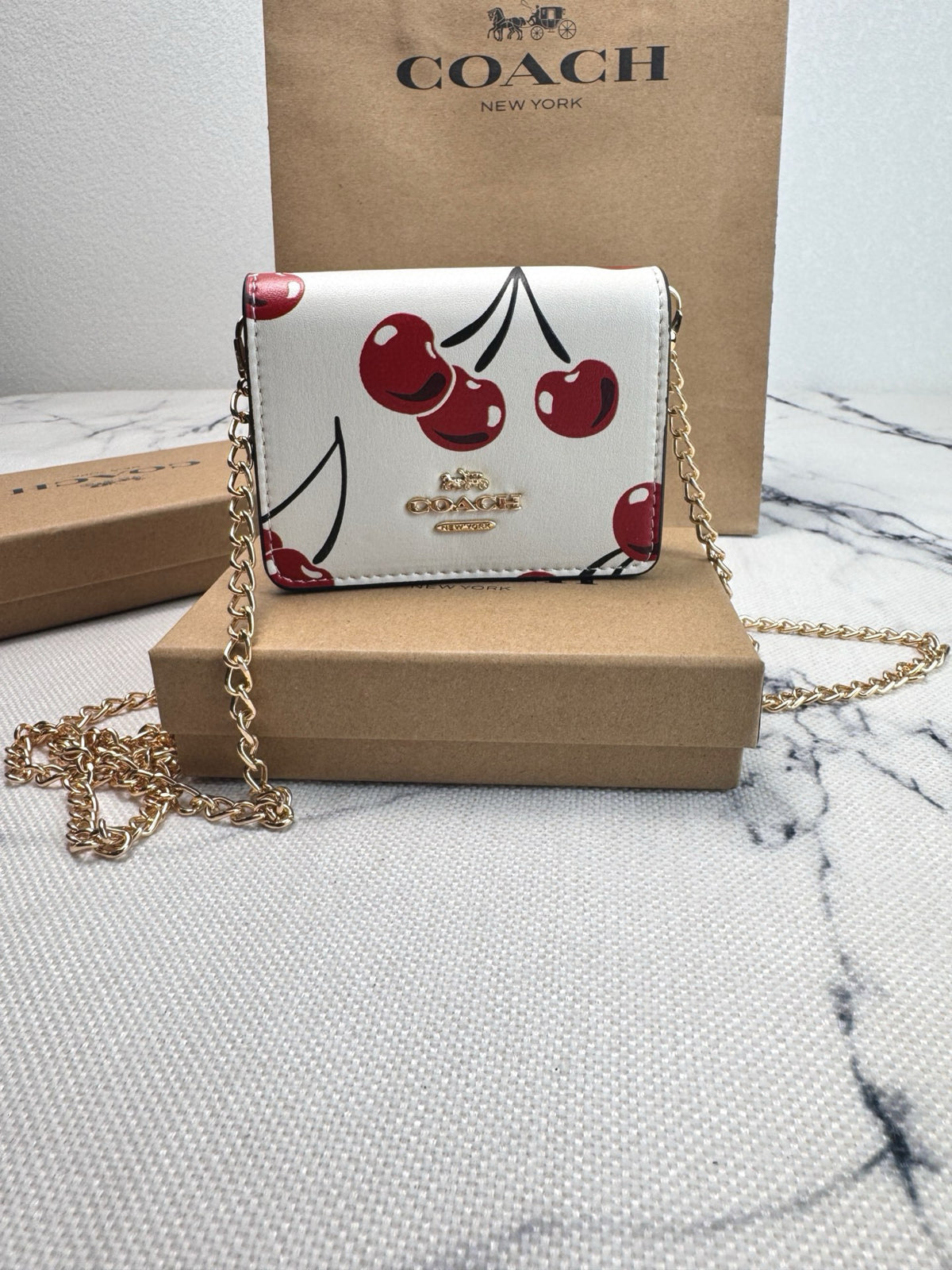 A Coach crossbody bag with a white base and a red cherry print, displayed on a brown cardboard box with its signature Coach box and paper bag in the background.