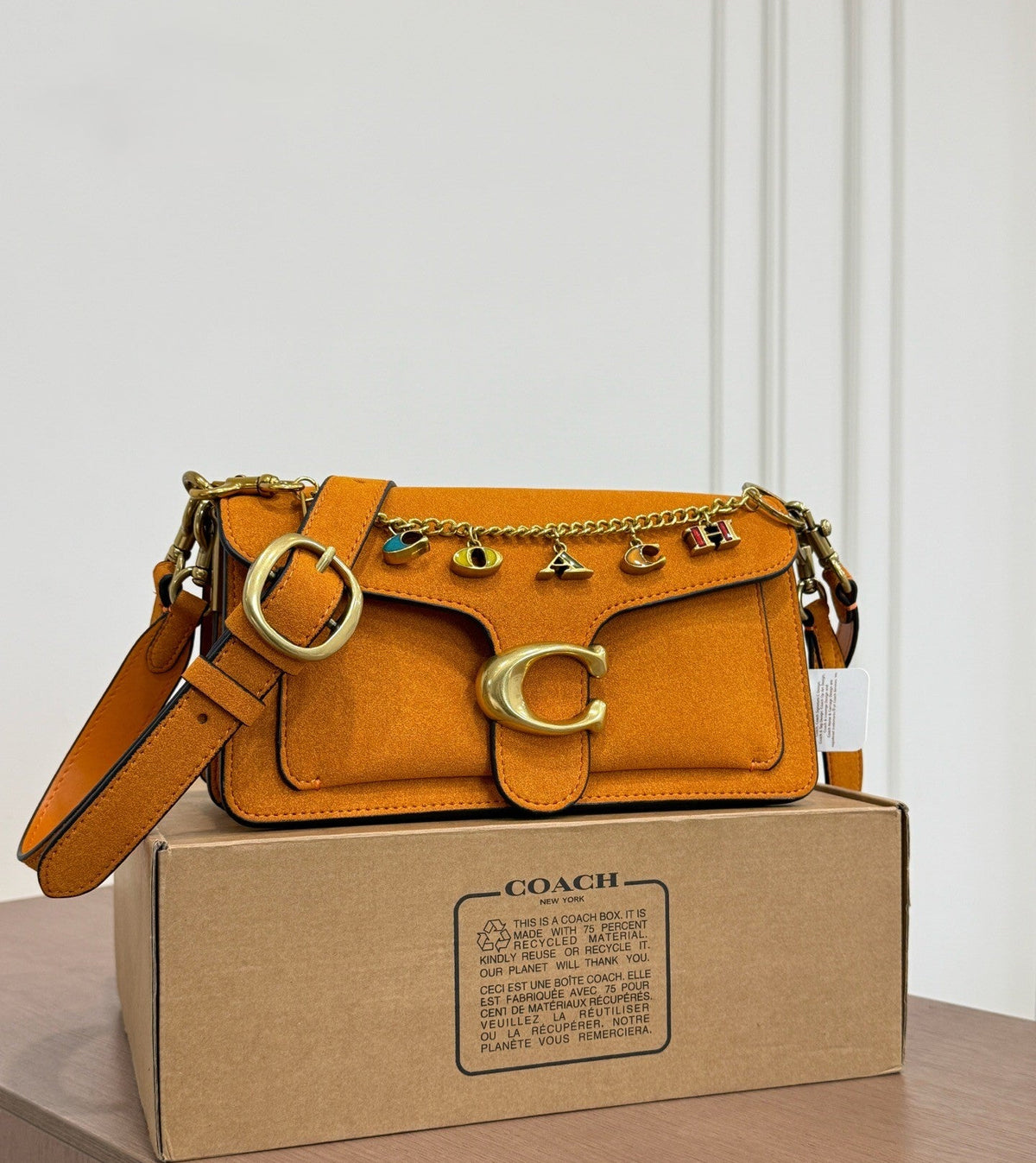 A Coach shoulder bag in a warm camel tan color, displayed on top of its original cardboard box. The bag features a prominent gold-tone 'C' logo on the front, a chain strap with 'COACH' letter charms, and a matching shoulder strap.