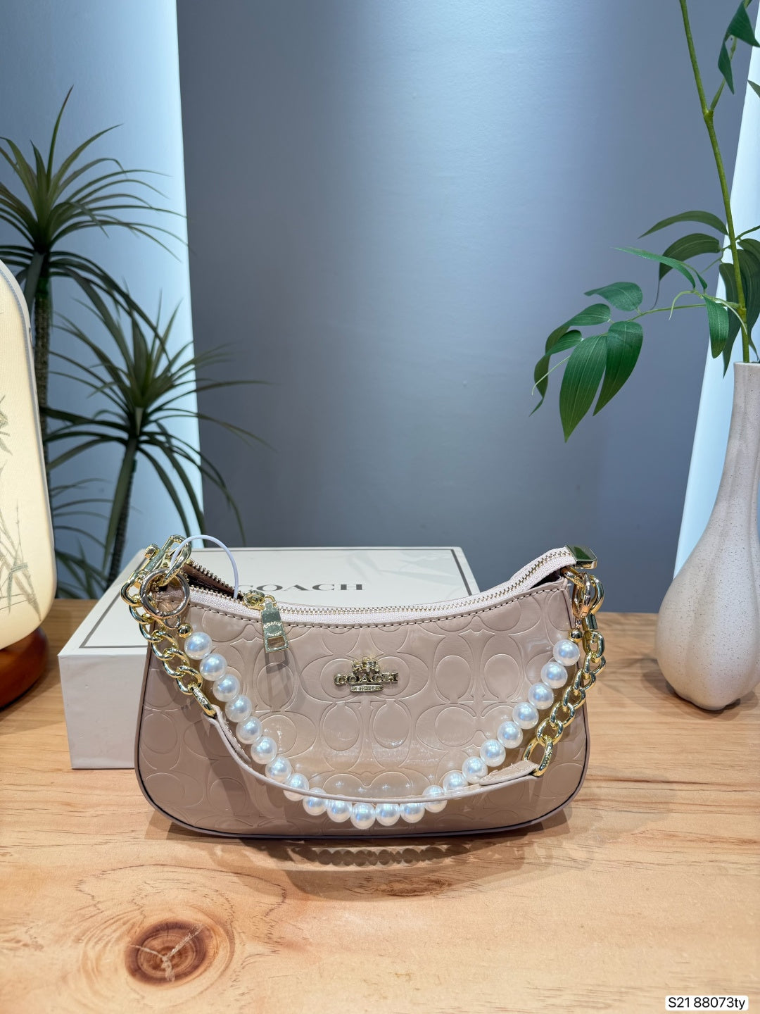 A Coach crescent-shaped shoulder bag in beige signature-coaster leather with a pearl bead strap and gold-tone chain accents, displayed on a wooden table with its box in the background.