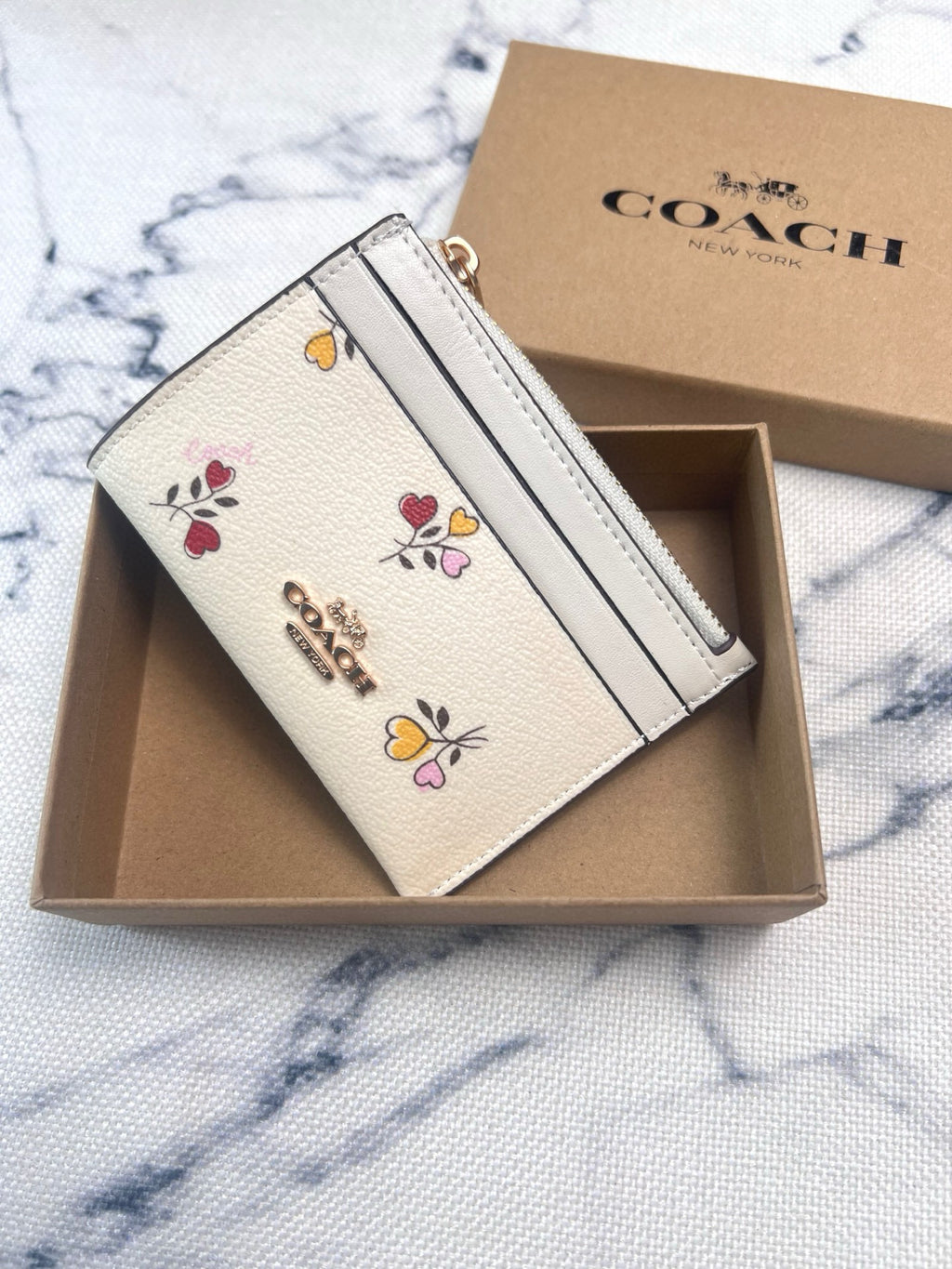 A Coach card holder wallet in cream-colored textured leather with colorful floral embroidery and a gold-tone logo, presented in its open brown box with the branded box lid behind it on a white marble surface.