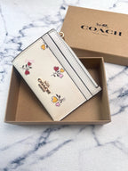 A Coach card holder wallet in cream-colored textured leather with colorful floral embroidery and a gold-tone logo, presented in its open brown box with the branded box lid behind it on a white marble surface.
