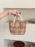Small woven straw tote bag with white handles and a tan leather logo patch. The bag has 'COACH' printed across the front in black.