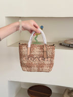 Small woven straw tote bag with white handles and a tan leather logo patch. The bag has 'COACH' printed across the front in black.